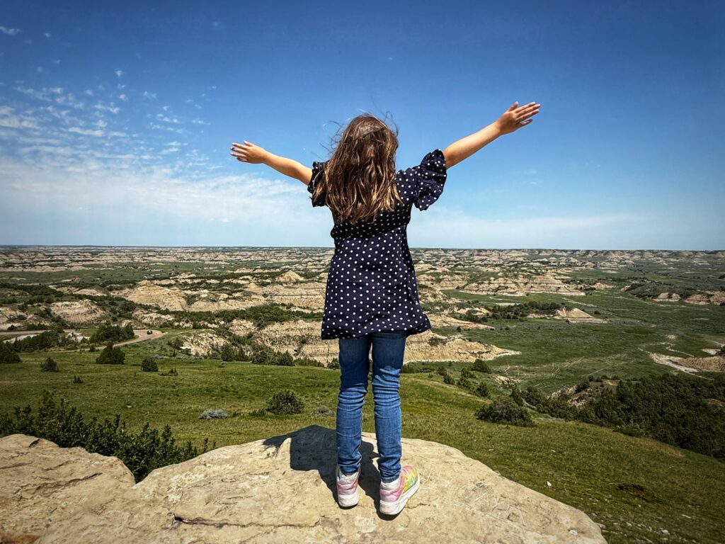 My daughter standing at the top of Buck Hill in Theodore Roosevelt National Park with her arms up 