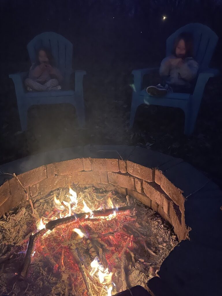 Two kids sitting in blue Adirondack chairs around a glowing backyard fire pit, enjoying one of the coziest fall outdoor activities for kids.