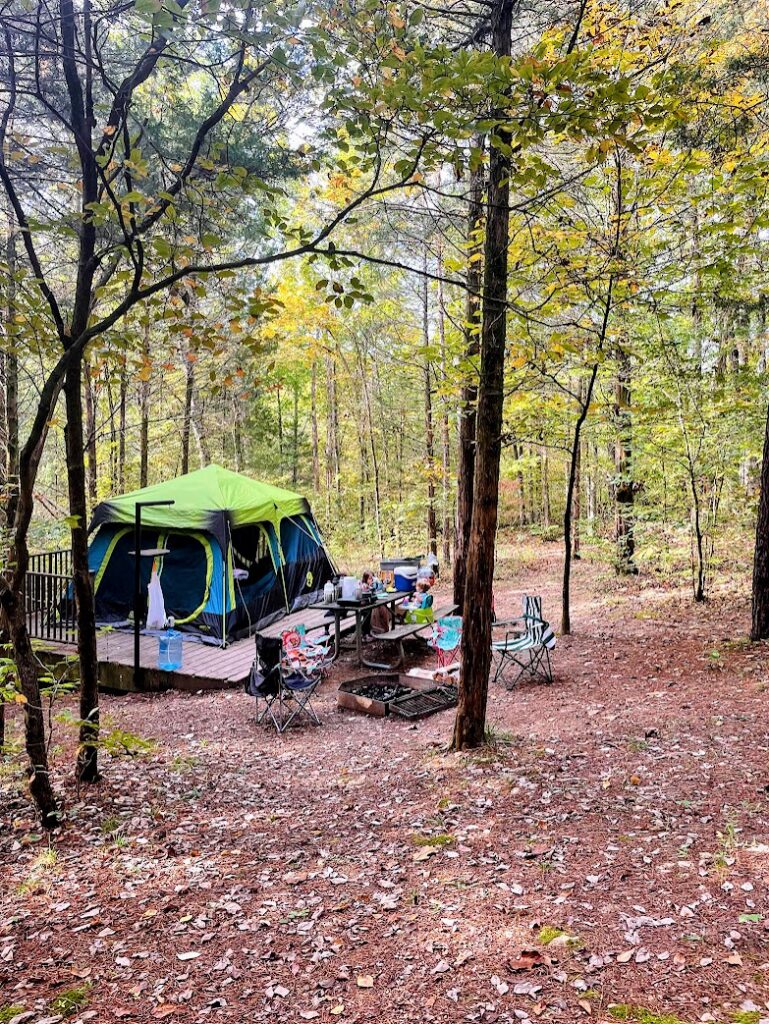 Family campsite set up with a green tent, picnic table, and chairs in a wooded area, showing camping as one of the best fall outdoor activities for kids.
