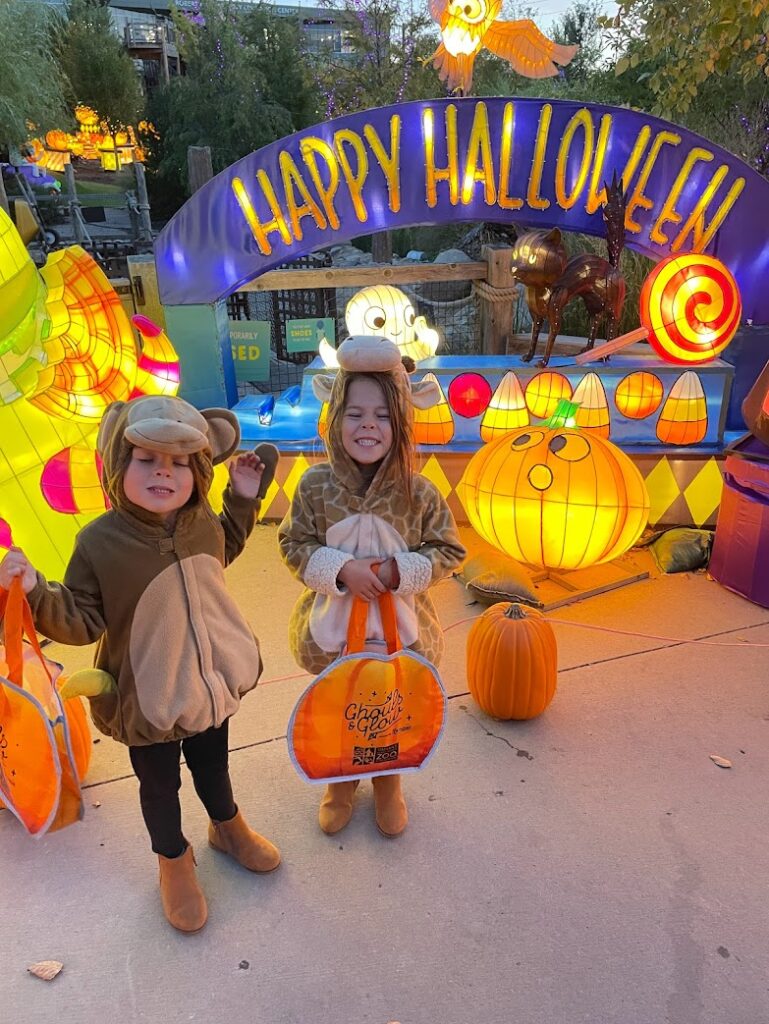 Kids in costumes holding trick-or-treat bags at a fall festival with Happy Halloween decorations. Fall festivals are lively fall outdoor activities for kids.
