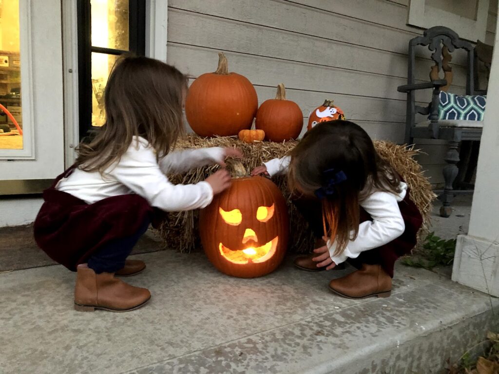 Two young girls admiring a glowing jack-o-lantern on a porch in fall. Carving pumpkins is one of the classic fall outdoor activities for kids.