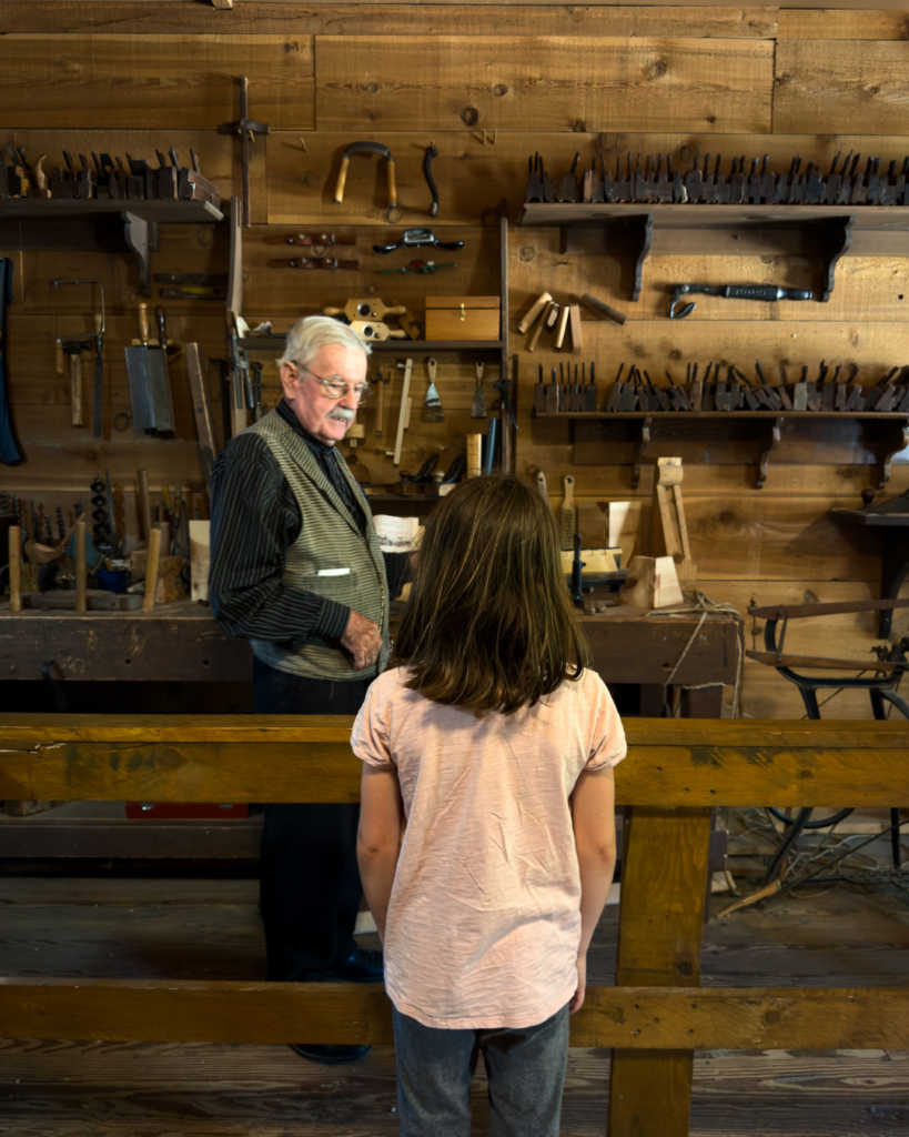 A child watches a historical interpreter explain tools and trades inside a recreated nineteenth-century woodworking and blacksmith shop.