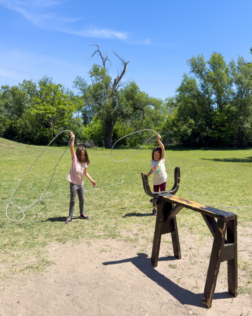 Two children practice lasso skills outdoors at an Old West attraction, learning cowboy techniques through hands-on family activities.