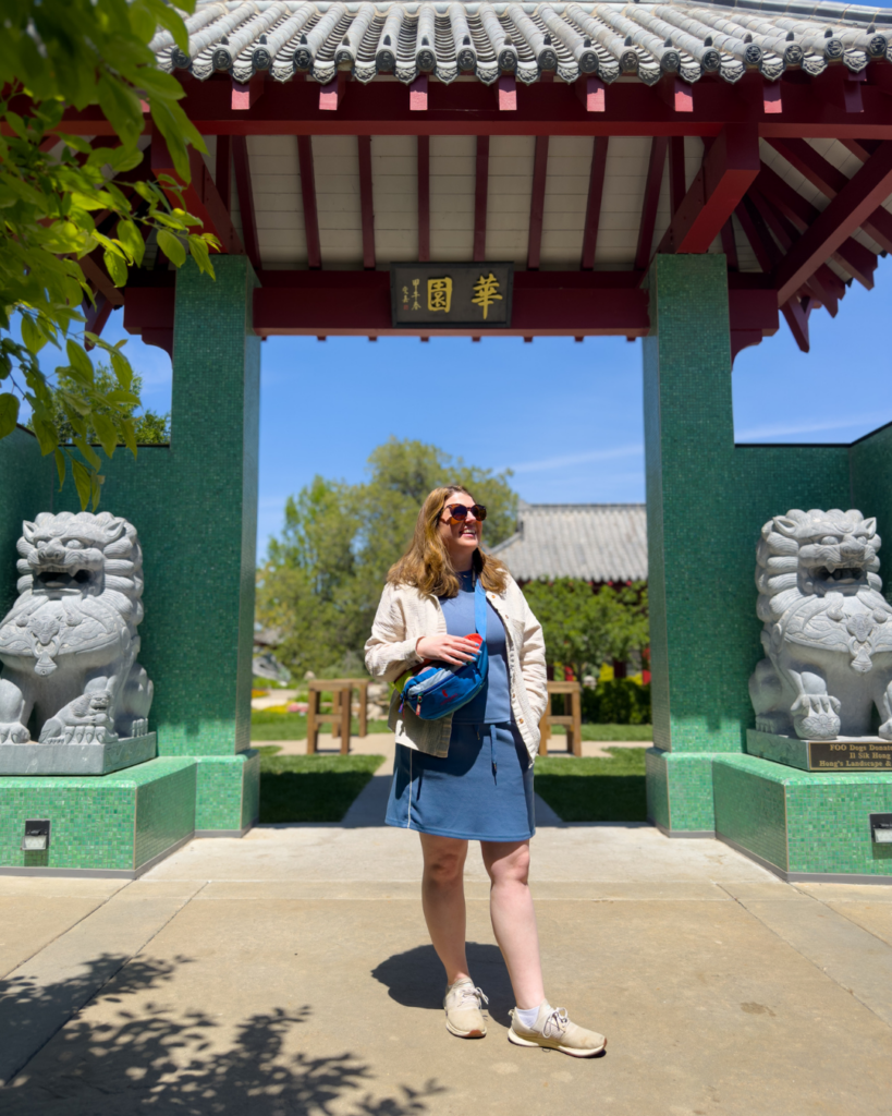 A visitor stands beneath a traditional Chinese garden gate flanked by stone guardian lions at a botanical garden in Wichita, Kansas.