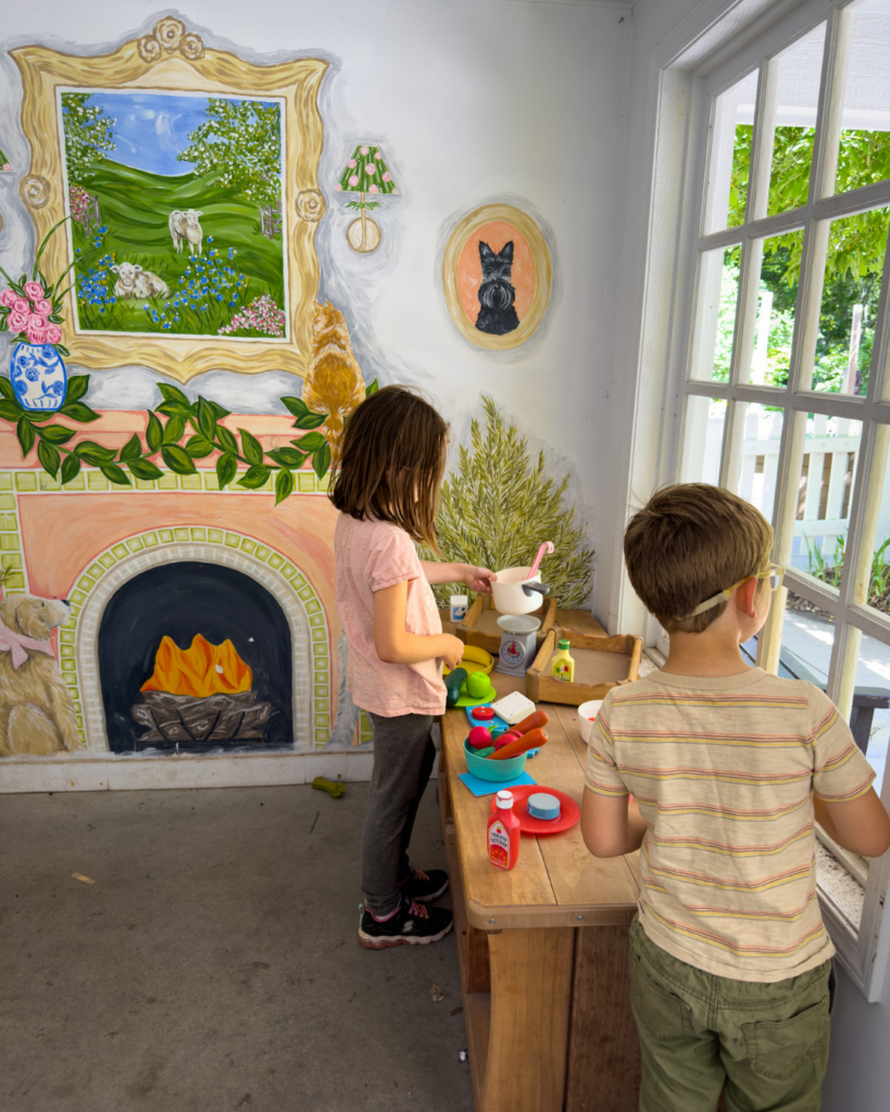 Two children play in a pretend kitchen inside an interactive children's garden, encouraging imaginative and hands-on learning.