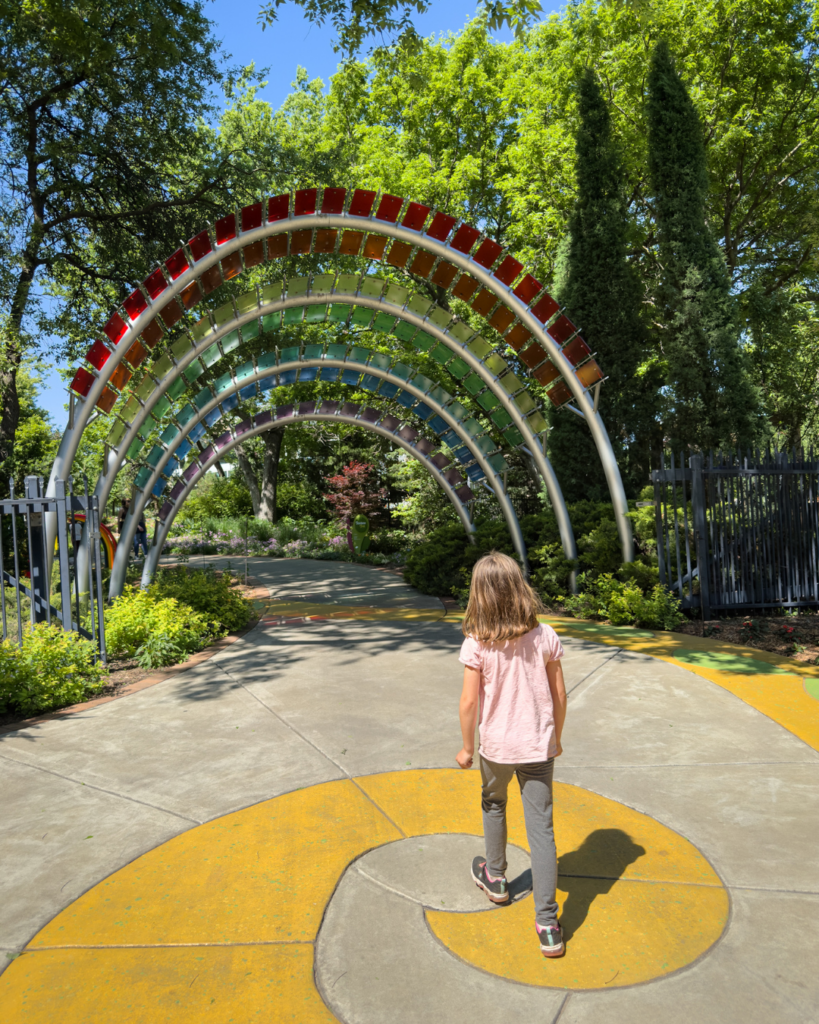 A child walks beneath a colorful rainbow archway surrounded by lush gardens and winding pathways at a botanical attraction.