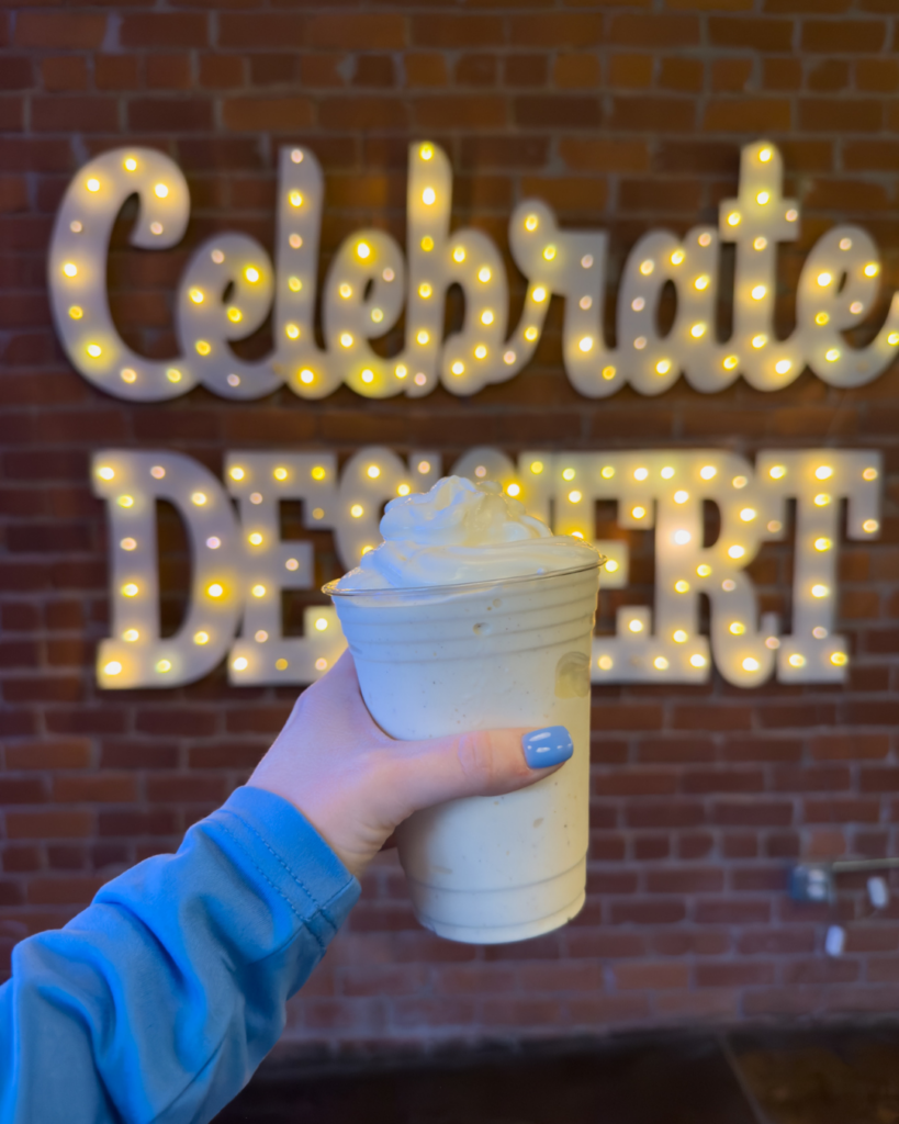 A creamy dessert shake topped with whipped cream is held in front of a glowing Celebrate Dessert sign in downtown Wichita.