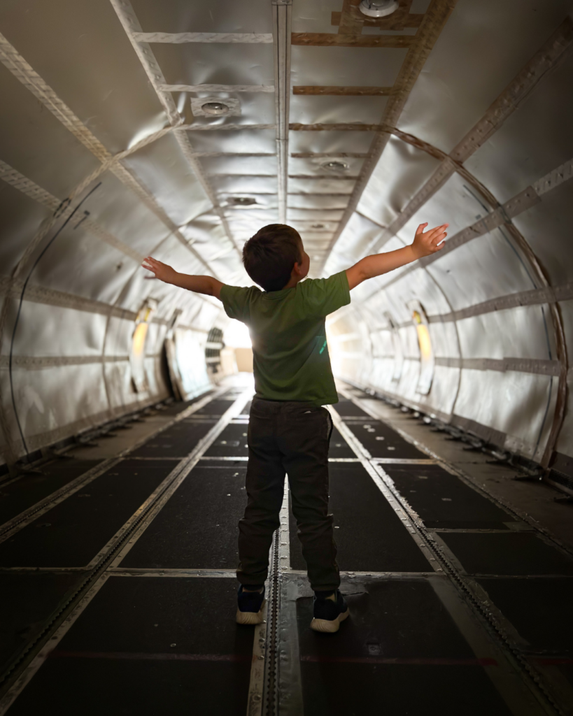 A young child stands inside a cargo airplane with arms outstretched, exploring the massive interior of a retired aircraft.