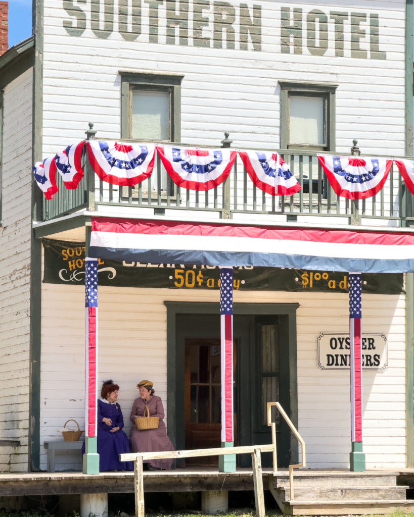 Two women in period clothing sit on the porch of the historic Southern Hotel, recreating daily life in a nineteenth-century frontier town.