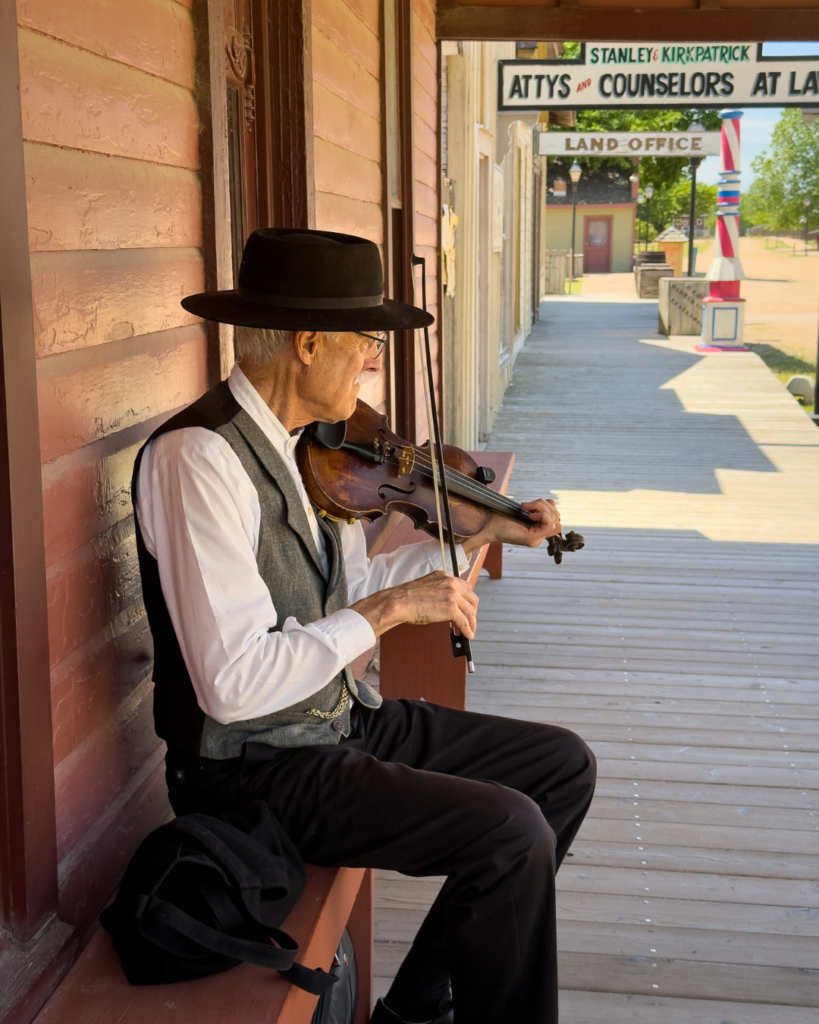 A fiddler dressed in period clothing performs on a wooden boardwalk, adding authentic music to a historic Old West town setting.